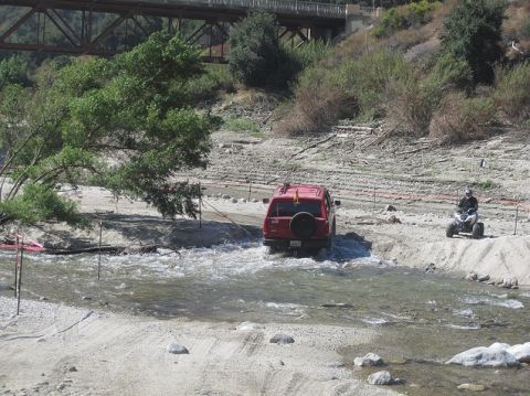 1988 Ford Bronco - Red SUV navigates a water crossing with an ATV nearby. Bridge visible in the distance.