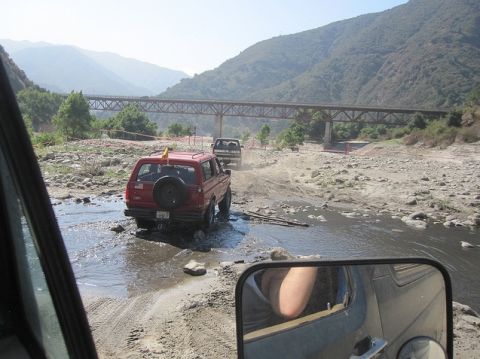 1988 Ford Bronco - Two vehicles navigate a dry riverbed with a large bridge overhead.
