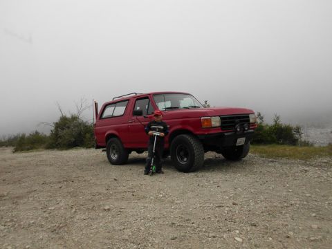 1988 Ford Bronco - A red Ford Bronco, likely from the late 1980s, is parked on a dirt road with a child and scooter beside it.