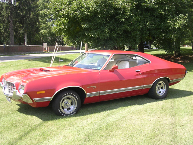 Red Ford Gran Torino Sport fastback with hood scoop and mag wheels parked on grass, driver's side view