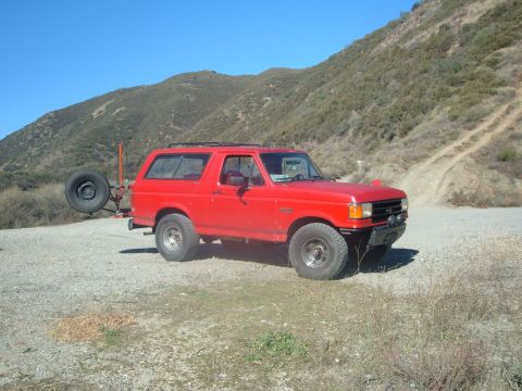 1988 Ford Bronco - A red Ford Bronco is parked on a dirt road with a spare tire mounted on the rear carrier.
