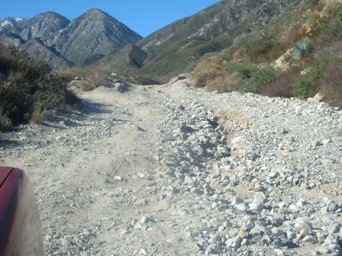 1988 Ford Bronco - A red vehicle fender is visible on a rocky dirt road winding through a mountainous landscape.