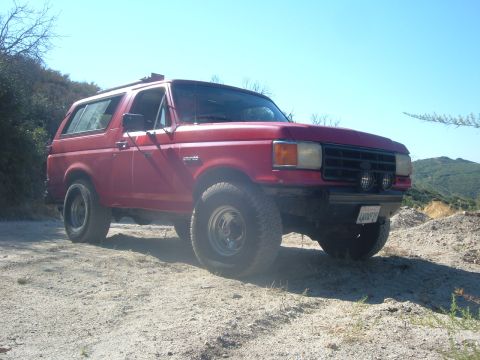 1988 Ford Bronco - This red 1988 Ford Bronco is parked on a dirt road with hills in the background.