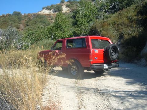 1988 Ford Bronco - Red Ford Bronco parked on a dirt road with dry grass and hills in the background.