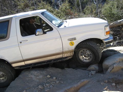 1996 Ford Bronco - A white Ford Bronco is shown off-roading over large rocks on a trail.