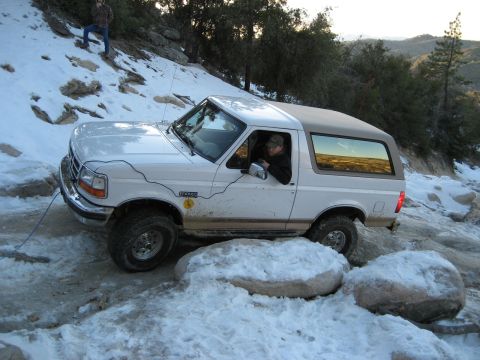 1996 Ford Bronco - A white 1996 Ford Bronco tackles a challenging off-road path covered in snow and rocks.
