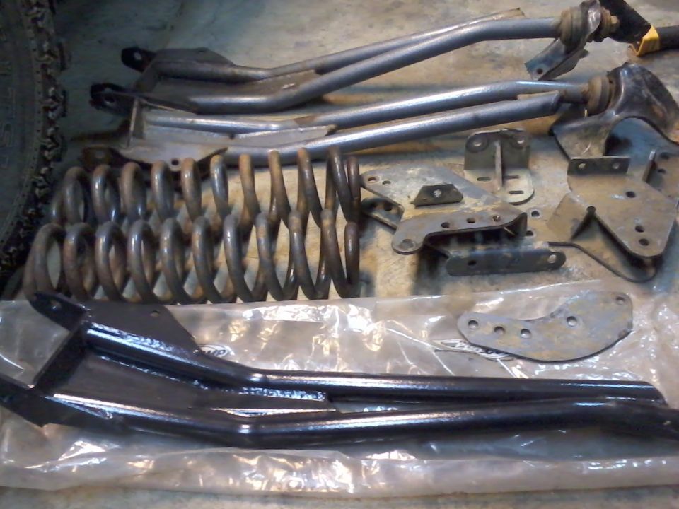 Several suspension components, including coil springs and control arms, laid out on a concrete floor. Some parts are black, others are gray and rusty