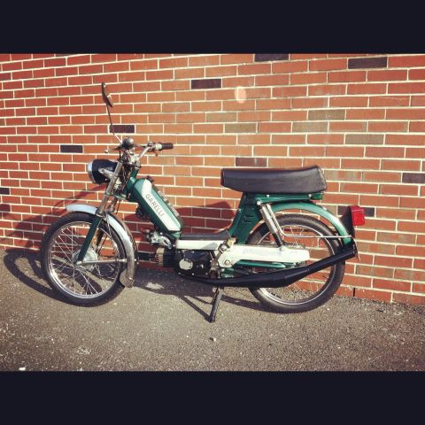 1981 Garelli Rally Sport - A green Garelli moped is parked on asphalt in front of a brick wall. The moped has a black seat and exhaust pipe.