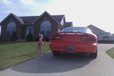 1999 Chevrolet Camaro - A red 1999 Chevrolet Camaro is parked in a driveway with a young girl nearby and a white truck in the background.