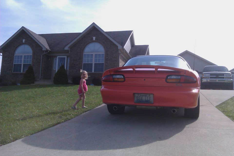 Red 1999 Chevrolet Camaro parked in driveway, rear view. A young girl stands on the grass. A white truck is parked behind it