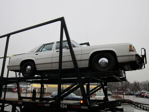 1991 Ford Crown Victoria LTD - A white 1991 Ford Crown Victoria LTD is transported on a car carrier, revealing its side profile and some front-end damage.