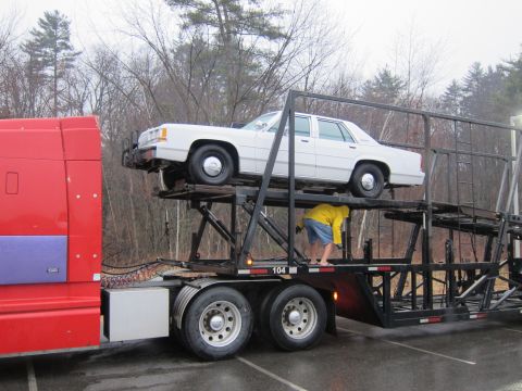 1991 Ford LTD Crown Victoria - A white sedan is being transported on a car carrier trailer, with a person in a yellow jacket attending to it.