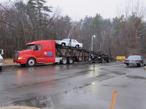 1991 Ford LTD Crown Victoria - A red car carrier transports a white 1991 Ford Crown Victoria LTD, while another car exits the scene.