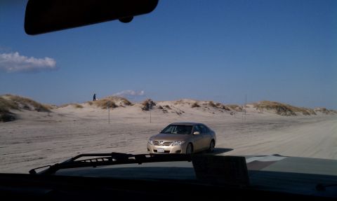 1979 Ford Bronco - A gold sedan is seen on a sandy beach, with dunes and a clear blue sky in the background.