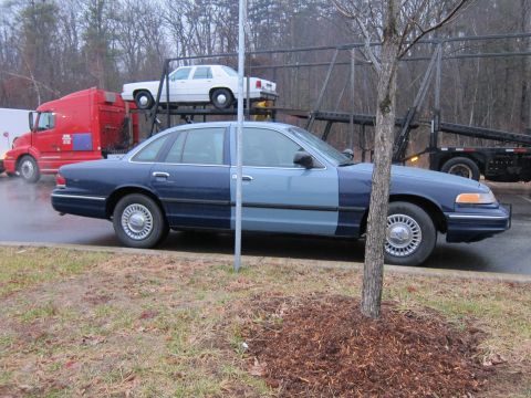 1991 Ford LTD Crown Victoria - A two-tone blue sedan, possibly a police car, is parked on the side of the road with a car carrier and semi-truck behind it.