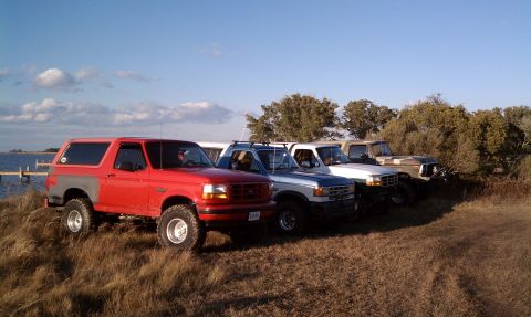 1979 Ford Bronco - Several SUVs and trucks are parked in a grassy area near the water under a blue sky.