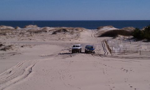 1979 Ford Bronco - Two SUVs navigate sandy terrain near the ocean, with dunes and a wooden fence visible.