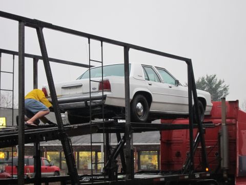 1991 Ford LTD Crown Victoria - A person is securing a white sedan on a car carrier, with another red vehicle visible below.