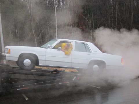 1991 Ford LTD Crown Victoria - A white sedan is being loaded onto a trailer, with a person in a yellow jacket inside and smoke billowing around the vehicle.