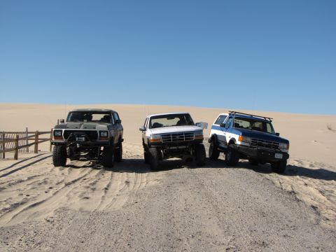 1979 Ford Bronco - Three SUVs are parked on a sandy terrain under a clear blue sky.