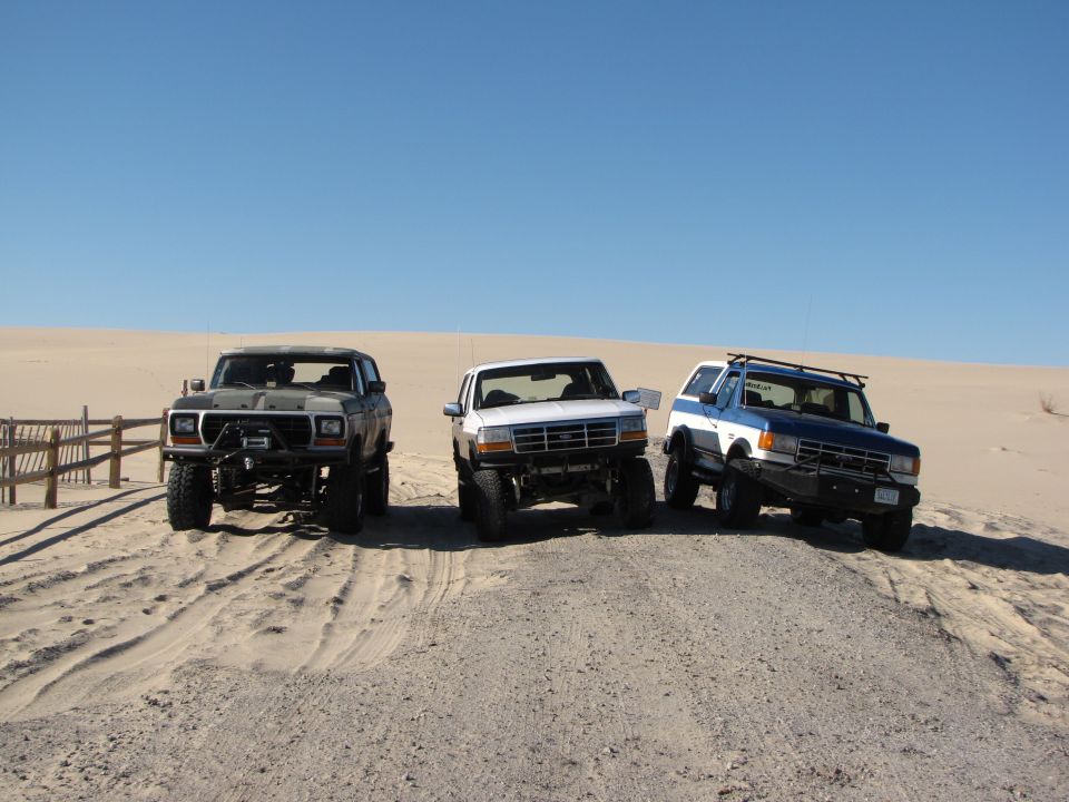 Three modified SUVs parked on sand, one dark gray, one white, and one blue and white. The dark gray SUV on the left appears to be a 1979 Ford Bronco