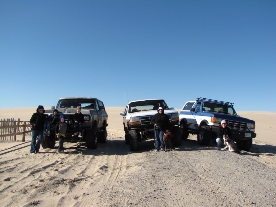 Three lifted SUVs on a sandy beach, one is a blue and white Ford Bronco