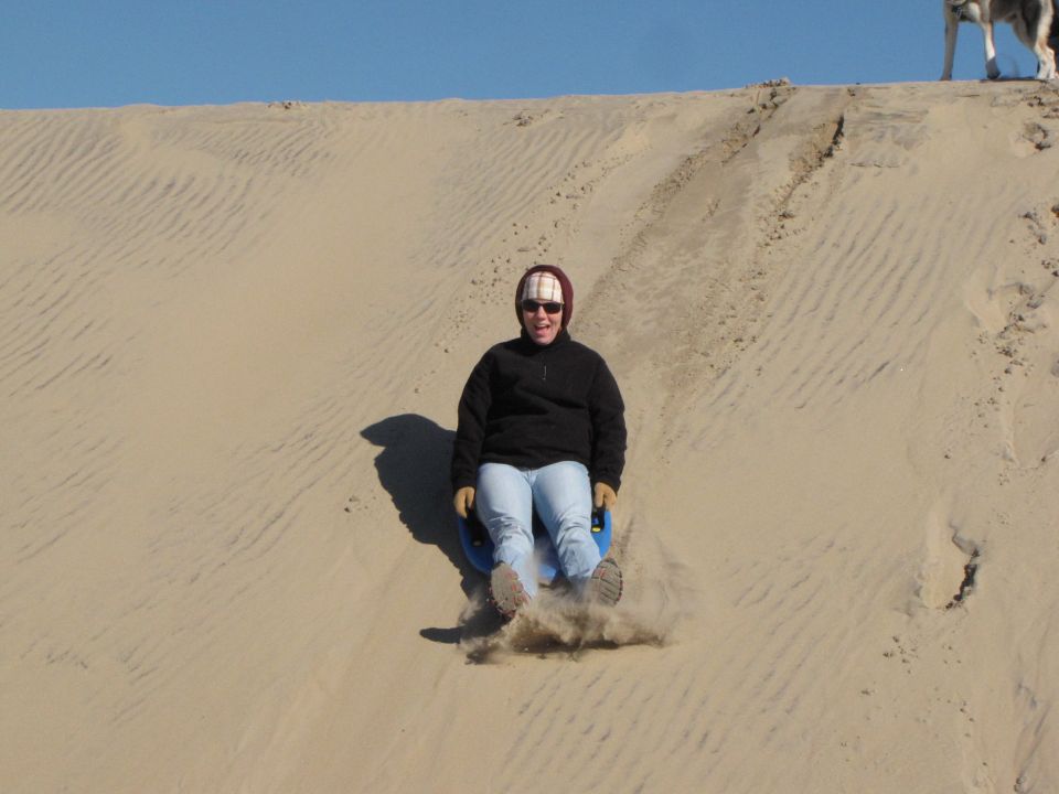 Person in black jacket and jeans sledding down a sand dune on a blue disc, kicking up sand