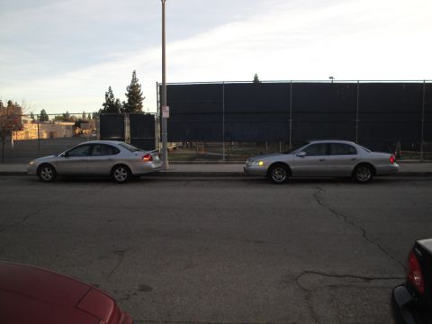 2000 Lincoln Continental - Two silver sedans, one a 2000 Lincoln Continental, are parked on a street next to a fence.