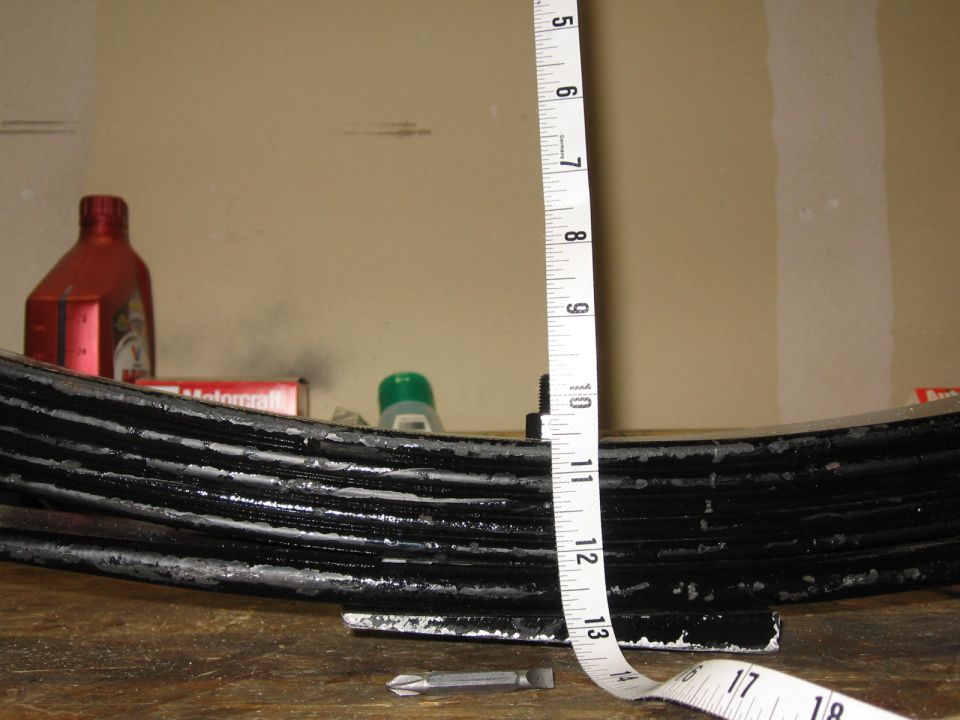 Black multi-leaf spring being measured with a tape at about 12 inches of arch on a workbench