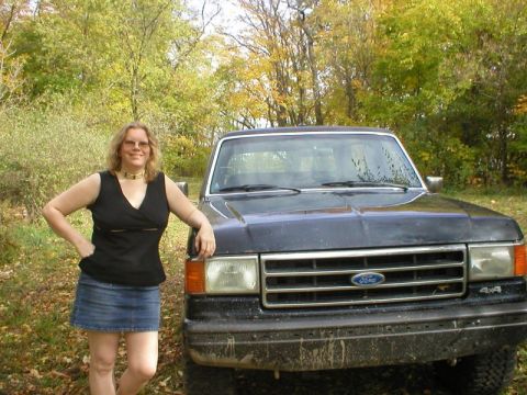 1989 Ford Bronco - Close front view of a dark blue Ford truck on a leaf-strewn trail in fall woods, with a person in a black top and denim skirt leaning again…