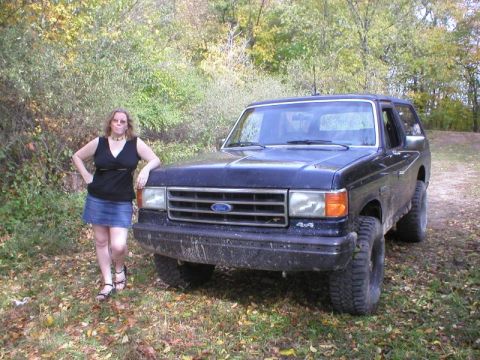 1989 Ford Bronco - Front three-quarter view of a dirty dark blue late-80s Ford Bronco parked on a leaf-strewn grass clearing beside autumn woods, with a perso…