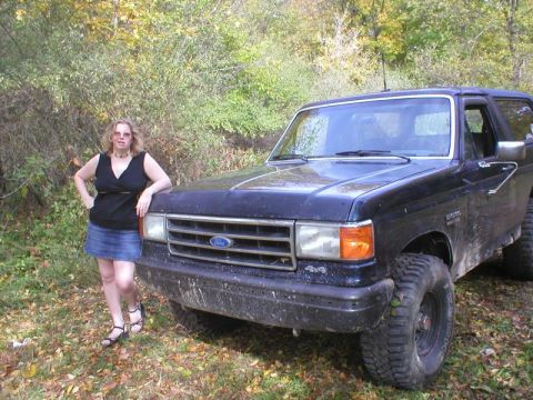 1989 Ford Bronco - A mud-spattered dark blue late-80s Ford Bronco parked on grass at a wooded treeline, shot from the front three-quarter with a person standi…