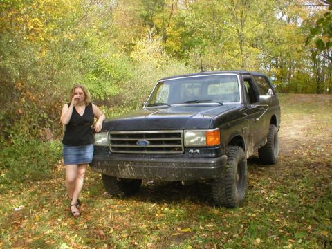 1989 Ford Bronco - Wide front three-quarter view of a lifted dark blue Ford Bronco with a removable hardtop, parked on a leaf-covered two-track through autumn…