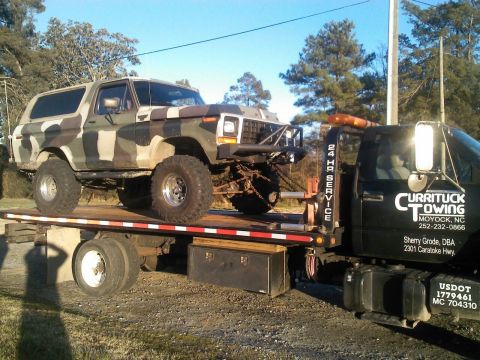 1979 Ford Bronco - A lifted, camouflage-painted SUV is loaded onto a flatbed tow truck with '24 HR SERVICE' visible on the side.