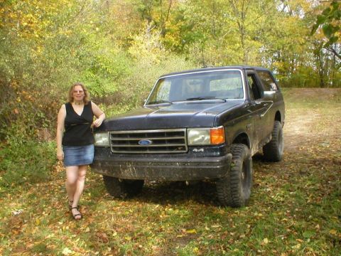 1989 Ford Bronco - A dark blue late-80s Ford Bronco parked beside a dirt two-track in autumn woods, seen from the front three-quarter with a person leaning on…