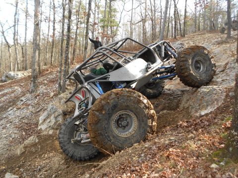 1967 Ford Mustang - Bare-aluminum tube-chassis rock buggy climbing a rocky off-camber ledge in leafless winter woods, shown from the driver's rear three-quarte…