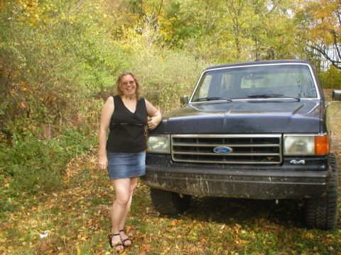 1989 Ford Bronco - Close-up front three-quarter view of a mud-splattered dark blue late-80s Ford Bronco parked on leaf-covered ground at a wooded edge, with a…