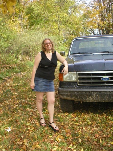 1989 Ford Bronco - Close front corner view of a dark blue late-80s Ford Bronco parked on leaf-covered grass in autumn woods, with a person leaning against the…