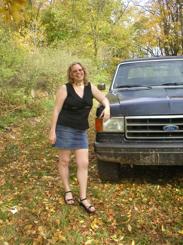 Person leaning on front fender of a dark blue late-80s Ford Bronco in autumn woods