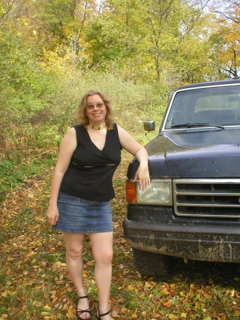 1989 Ford Bronco - A person in a black top and denim skirt stands beside the front fender of a dark blue Ford truck parked on a leaf-covered trail in autumn w…