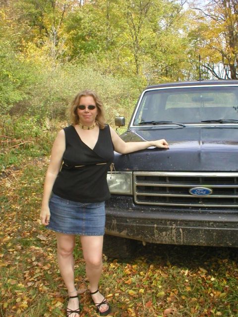 1989 Ford Bronco - A person in a black top and denim skirt rests a hand on the mud-flecked hood of a dark blue Ford truck parked on a leaf-covered trail in au…