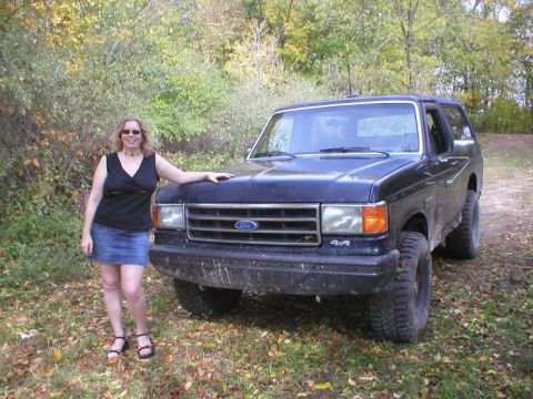 1989 Ford Bronco - A dark blue late-80s Ford Bronco parked at the edge of a dirt two-track in autumn woods, viewed from the front three-quarter with a person…