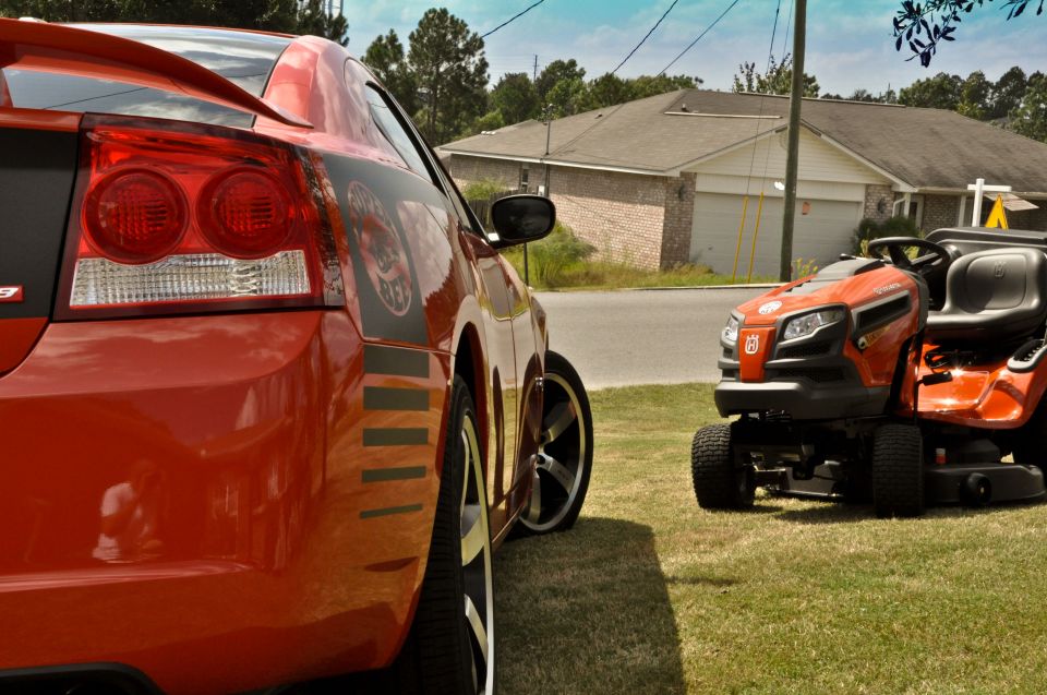 Red 2009 Dodge Charger rear quarter panel with black stripe and decal, parked on grass next to an orange riding mower