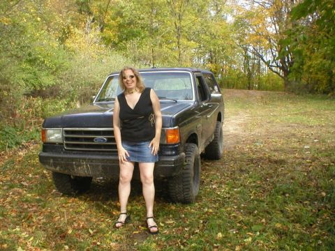 1989 Ford Bronco - A person in a black top and denim skirt stands in front of a lifted, mud-covered dark blue Ford Bronco on a grassy two-track through fall w…