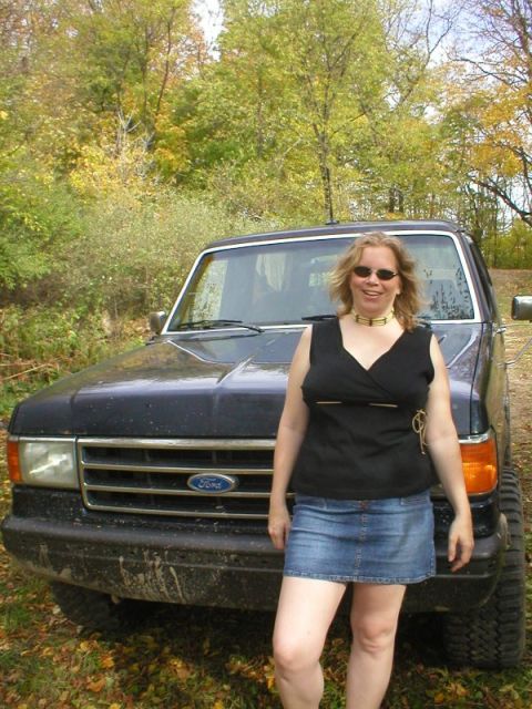 1989 Ford Bronco - A person in a black top and denim skirt stands directly in front of a mud-spattered dark blue Ford truck on a leaf-covered trail in fall wo…
