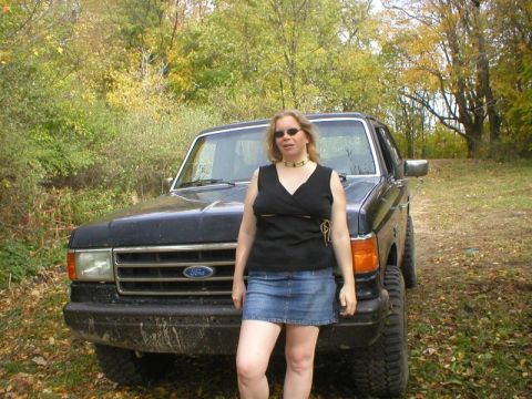 1989 Ford Bronco - A dark blue late-80s Ford Bronco parked on a dirt trail in fall woods, shot from the front with a person standing in front of the grille. M…
