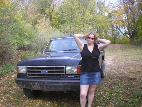1989 Ford Bronco - Front three-quarter view of a mud-splattered dark blue Ford Bronco parked on a leaf-covered two-track, with a person in a black top and den…