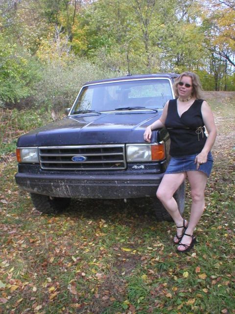 1989 Ford Bronco - A person in a black top and denim skirt leans against the front fender of a mud-spattered dark blue Ford Bronco parked on a grassy, leaf-st…