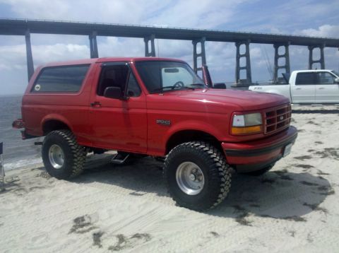 1995 Ford Bronco - Red 1995 Ford Bronco parked on a sandy beach with a bridge and another truck in the background.