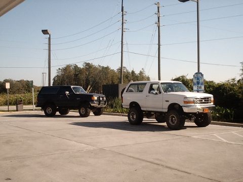 1996 Ford Bronco - Two lifted Ford Broncos, one white and one black, are parked side-by-side in a lot.